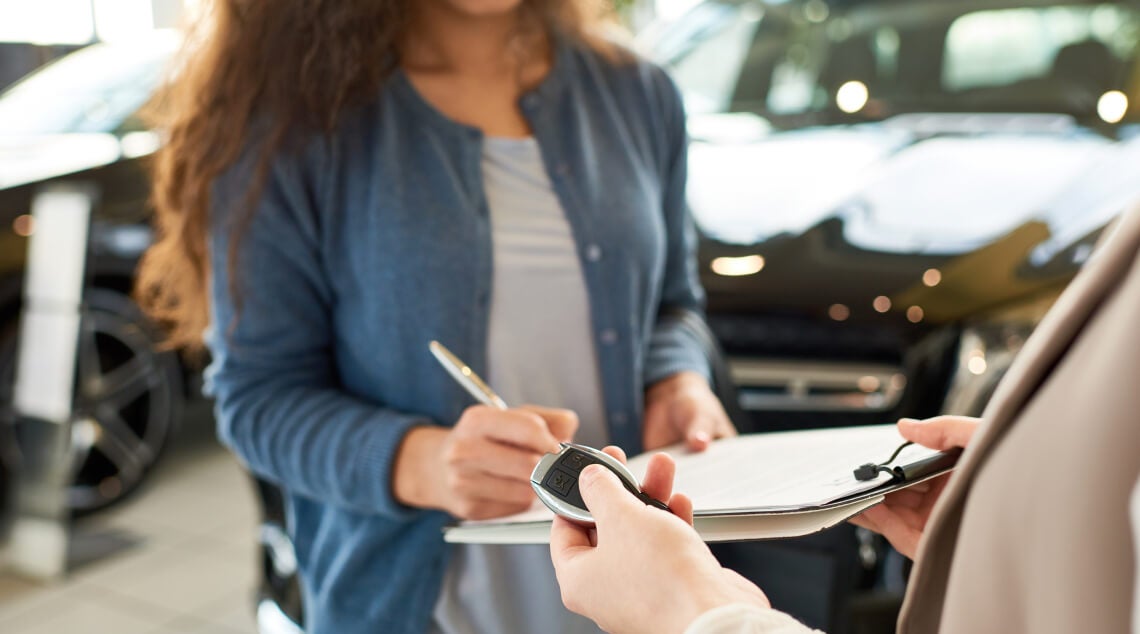 Customer signs dealership paperwork while receiving a vehicle key fob from a salesperson.
