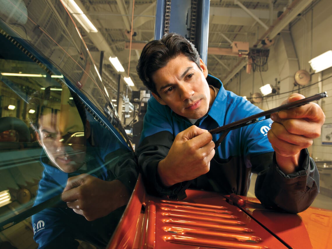 Mopar technician inspects a windshield wiper blade on a red vehicle in a dealership service bay.