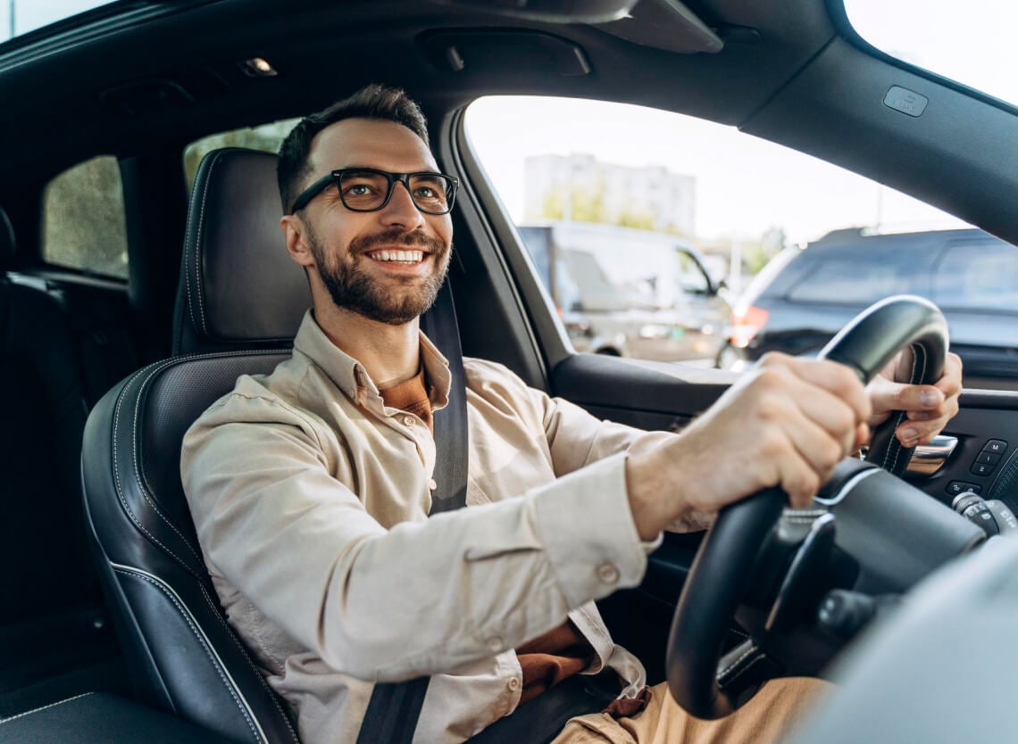 Smiling man wearing glasses drives in traffic, viewed from the front passenger seat.